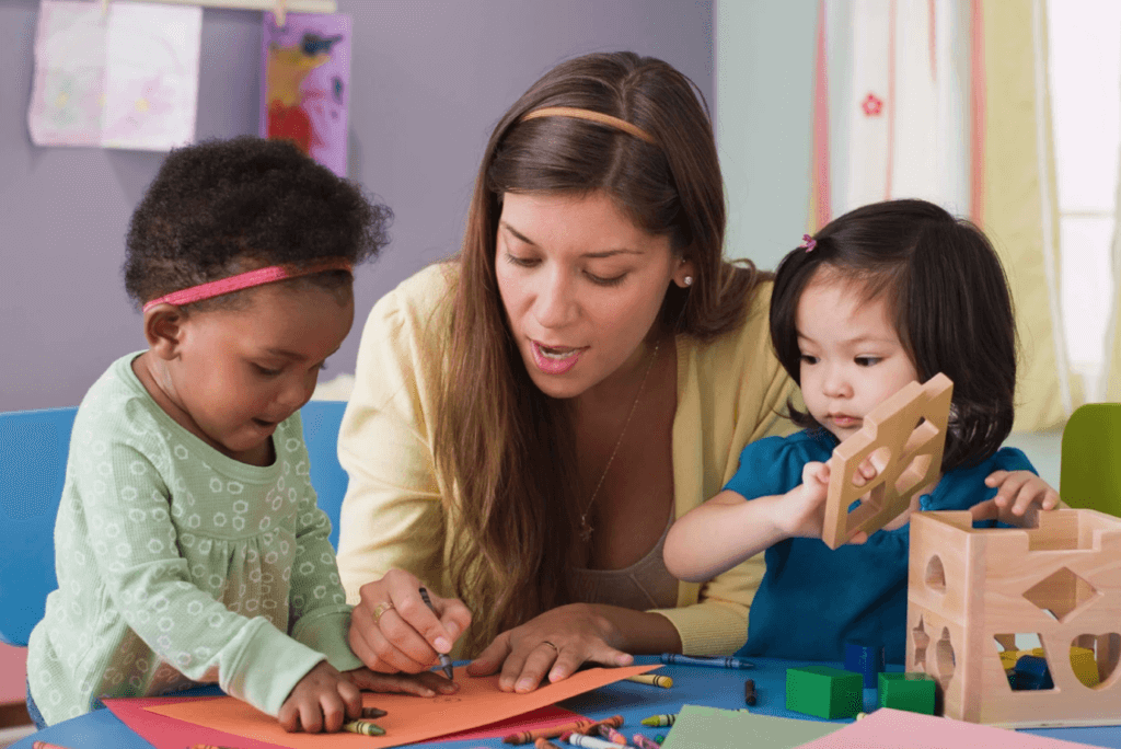 a woman holds a crayon as she shows a young girl how to draw with another young girl playing with wooden blocks next to her
