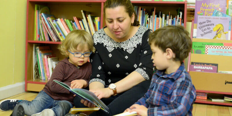 a woman reads a book to two young boys as they sit in front of a bookshelf in a French preschool in New York