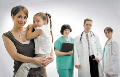Asian mother in tank top holds toddler girl in white dress; smiling nurse in scrubs with chart, male doctor in white coat with stethoscope, and female doctor stand behind on gray background.