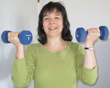 A smiling woman in a green shirt holds two blue 8-pound dumbbells while exercising against a plain white wall.