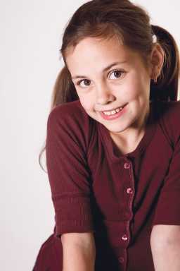 A young girl with brown hair in pigtails wearing a maroon cardigan, smiling and looking directly at the camera.