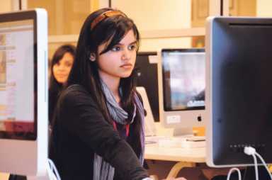 A young woman with long dark hair and a grey scarf looks intently at a computer monitor in a bright classroom or lab.