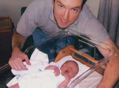 A man with blue eyes leans over a hospital bassinet looking at a yawning newborn baby wrapped in a white blanket.