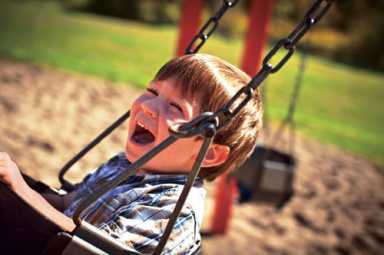 A young boy in a plaid shirt laughs with joy while playing on a swing set at a sunny outdoor playground.