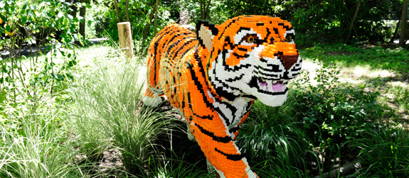 lego-safari A large, life-sized tiger sculpture made entirely of orange, black, and white LEGO bricks in a garden.