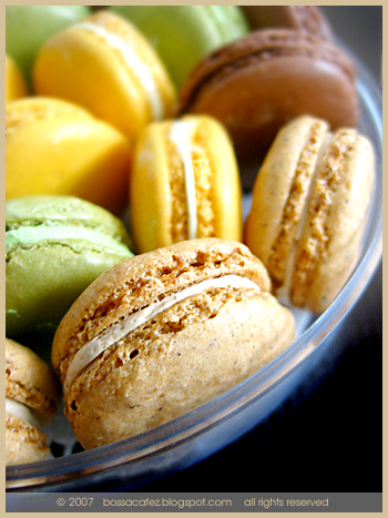 Assorted colorful macarons in yellow, green, and brown stacked together in a clear glass bowl.