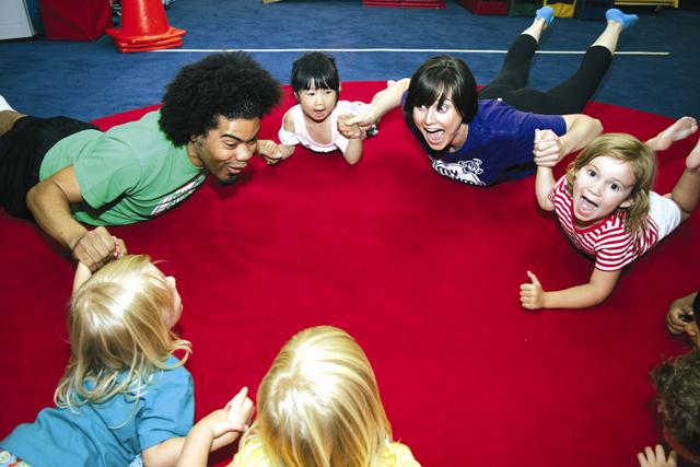 A circle of smiling children and two adults lie on their stomachs on a red circular mat, holding hands in a bright gym.