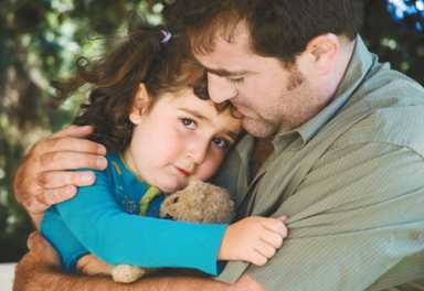A man tenderly hugging a young child who is holding a small stuffed animal.