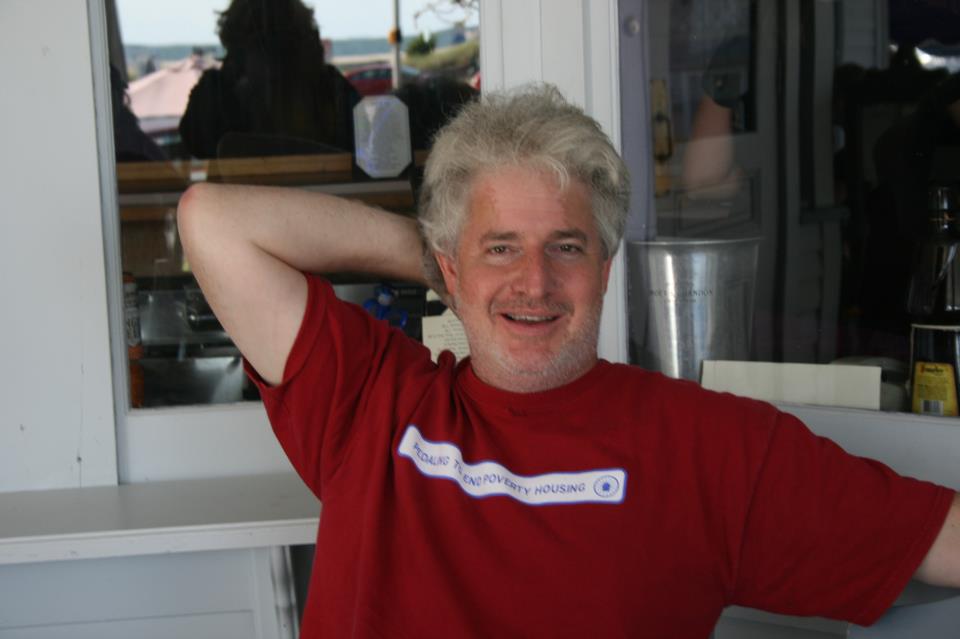 A man with graying hair wearing a red t-shirt smiles with one arm behind his head, sitting in an outdoor patio area.