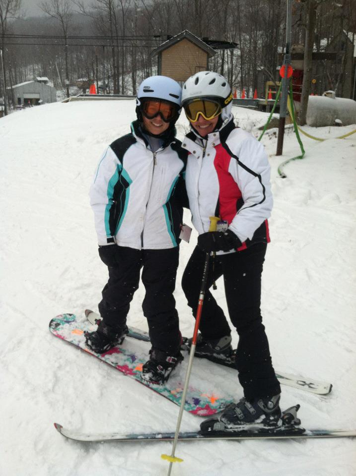 Two people in helmets and goggles smile while posing on a snowy slope with a snowboard and skis.