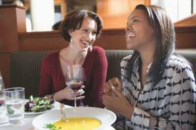 Two women smiling and laughing together while enjoying a meal and wine at a restaurant.