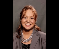 Portrait of a smiling woman with reddish-brown hair wearing a grey blazer and a pearl necklace.