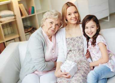 Three generations of women—grandmother, mother, and daughter—smiling together on a couch while holding a small gift.