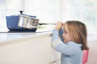 Young child reaching up toward a silver pot handle on a kitchen stove, illustrating a home safety hazard.
