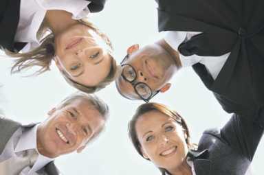 Low-angle view of four smiling professionals in business attire looking down in a huddle against a bright sky.