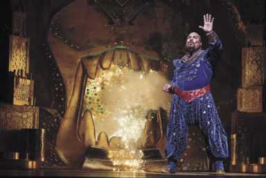 An actor in a sparkling blue genie costume stands on a stage next to a giant golden mouth prop emitting glowing light.