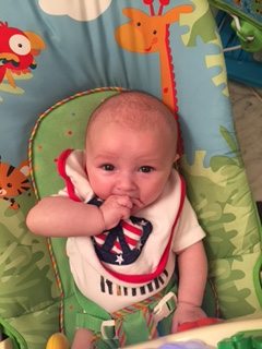 A baby with their thumb in their mouth sits in a colorful bouncer decorated with a giraffe and other animals.