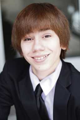 Headshot of a smiling boy with brown hair and bangs, wearing a black suit jacket, white shirt, and black tie.