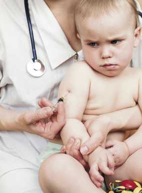 A healthcare worker with a stethoscope gives a vaccination to a nervous-looking baby sitting on someone's lap.