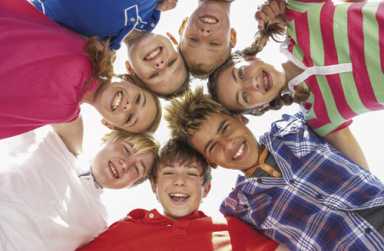 A group of diverse, smiling children huddle in a circle and look down at the camera against a bright sky.