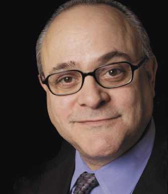 A close-up portrait of a man with glasses and a blue shirt smiling against a solid black background.