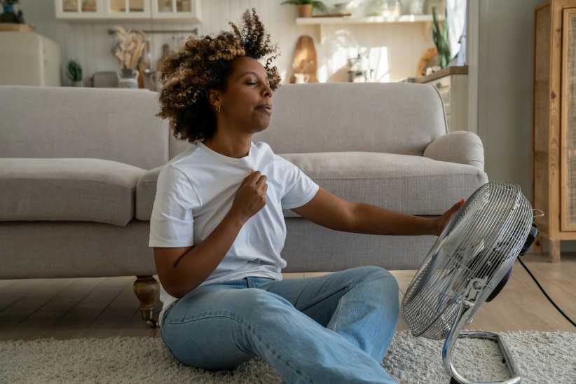 Young African American woman cooling down by ventilator at home, feeling unwell with high temperature during hot weather, sitting on floor in front of electric fan during extreme heatwave, menopause