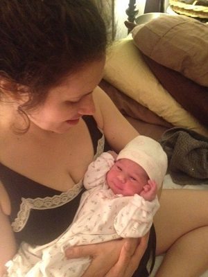 A woman in a black top gazes down at a newborn baby wearing a white cap and matching patterned onesie.