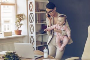 A woman in a blazer talks on a cell phone while holding a baby in an office setting near a laptop and baby bottle.