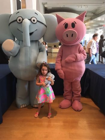 A young girl stands between a large elephant and a pink pig mascot, holding a small stuffed toy in a brightly lit room.