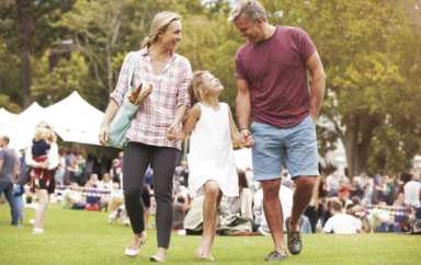 A mother and father hold hands with their daughter while walking across a grassy field at an outdoor event.