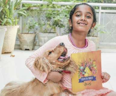 A young girl in a pink shirt sits on the ground, hugging a golden dog and holding a book titled 'Believe in Yourself'.