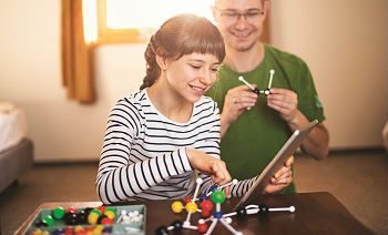 A girl and a man smiling while using a tablet and building molecular models with colorful kits.