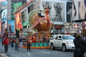 A giant turkey parade float in Times Square, surrounded by crowds, city billboards, and a white SUV in the foreground.