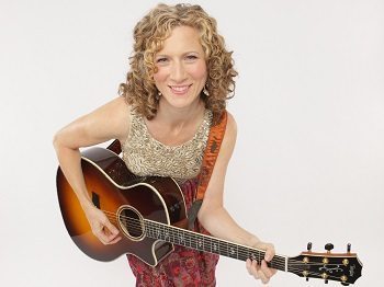 A woman with curly blonde hair smiles while playing an acoustic guitar against a plain white background.
