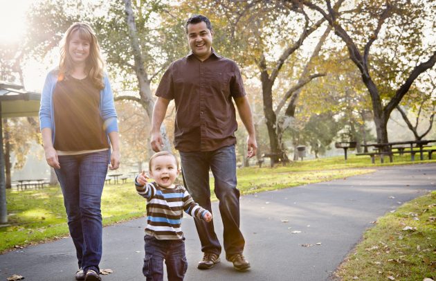 A smiling mother and father walk on a park path behind their toddler, who points forward with a joyful expression.