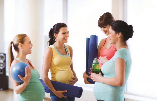 Four pregnant women in colorful workout clothes stand in a bright room, smiling and chatting while holding yoga mats and a blue exercise ball.
