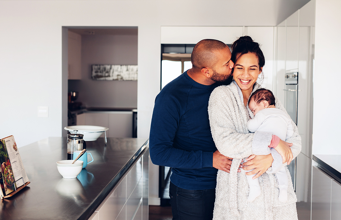 Man kissing his wife holding a newborn baby boy in kitchen. Lovely young family of three in morning in kitchen.