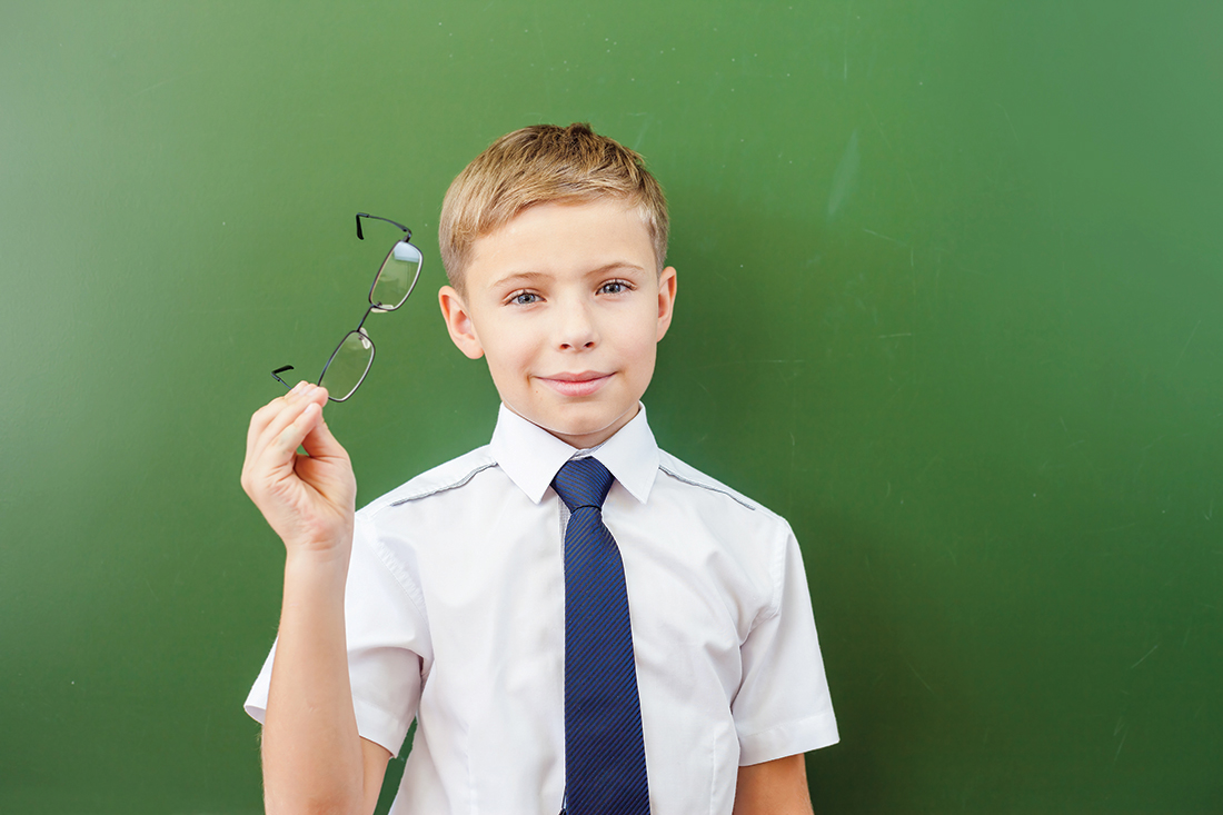 Successful schoolboy standing near the blackboard in a school classroom, dressed in a school uniform, and holding medical glasses. Boy's poor eyesight