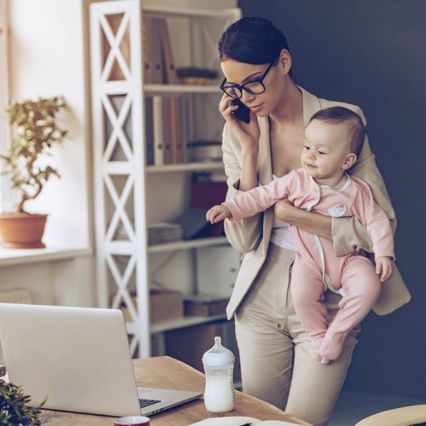 It is not easy to be a working mom! Young beautiful businesswoman talking on mobile phone and looking at laptop while standing with her baby girl at her working place
