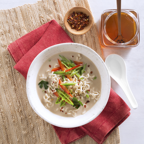 flatlay shot of ramen noodles in a white bowl on a wooden table with a red napkin and honey on the side as a garnish