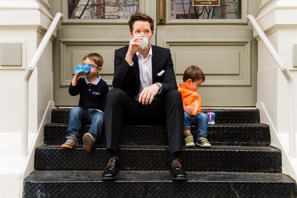 dad sitting on stoop with two young kids drinking from sippy cups