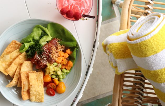 aerial viw of poke bowl, pink cocktail, and beach towel poolside at a fancy hotel