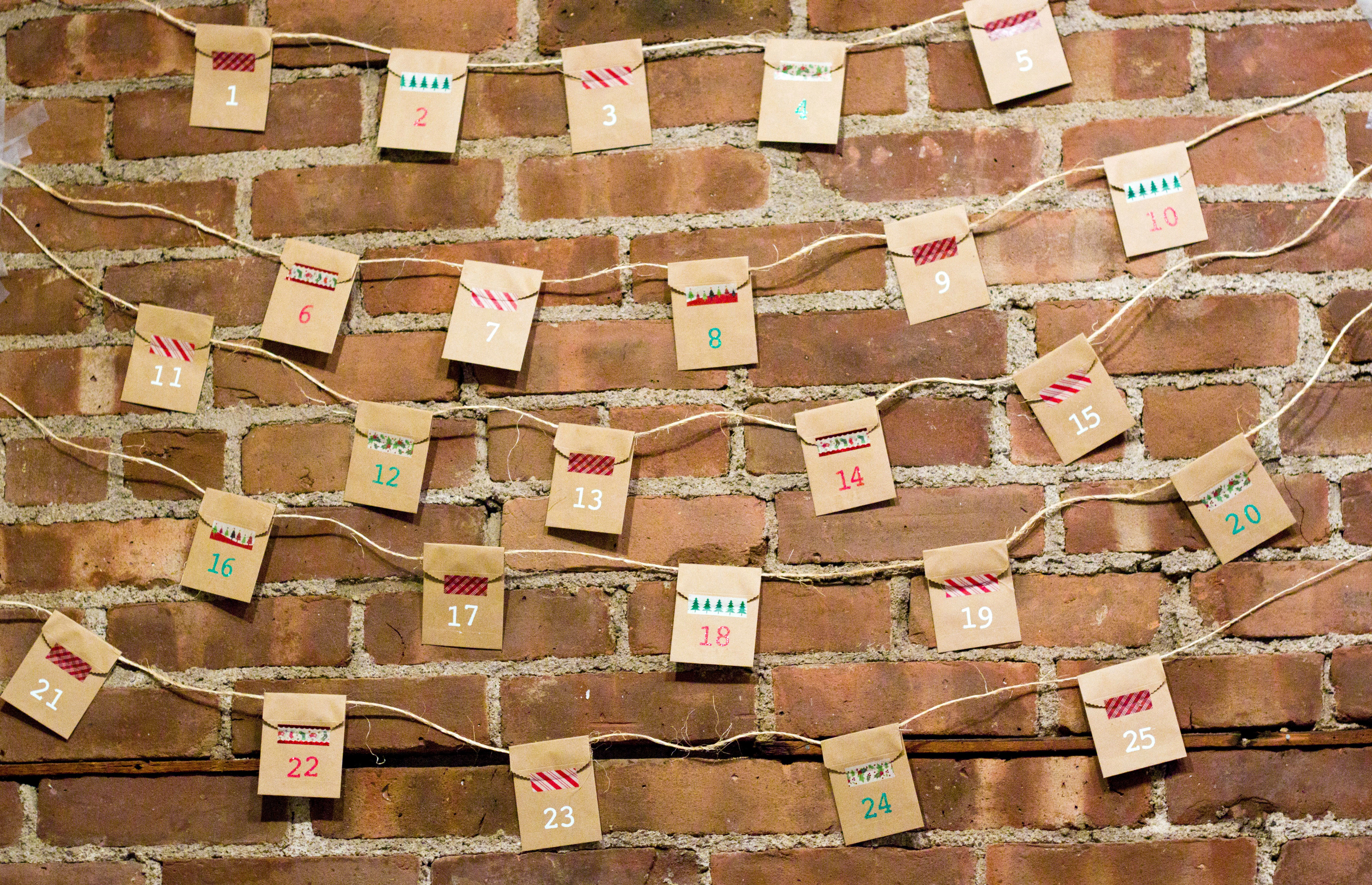 Small brown paper bags numbered for an advent calendar hang from twine against a red brick wall.