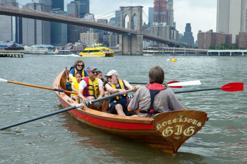 group of people paddling in rowboat with Brooklyn Bridge, water taxi and New York skyline in background