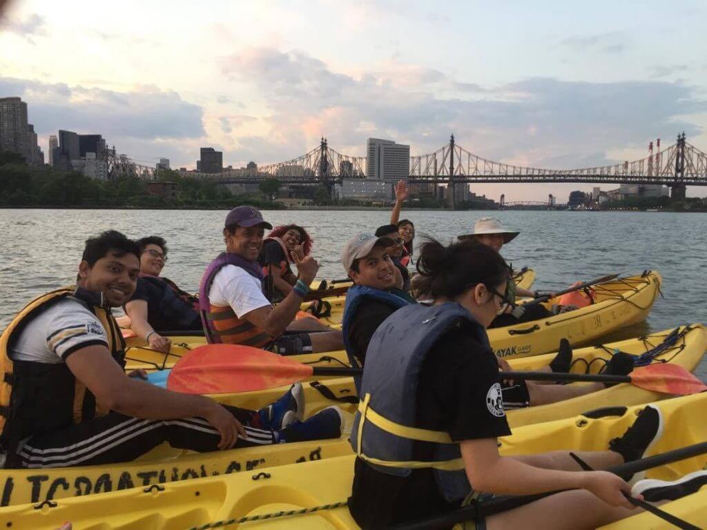 people in several yellow kayaks smile and give thumbs up with the Queensboro Bridge in the distance