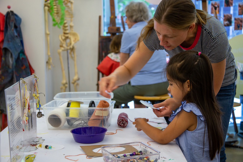 a woman and girl make necklaces with yarn and beads as other people stand near a skeleton in the background