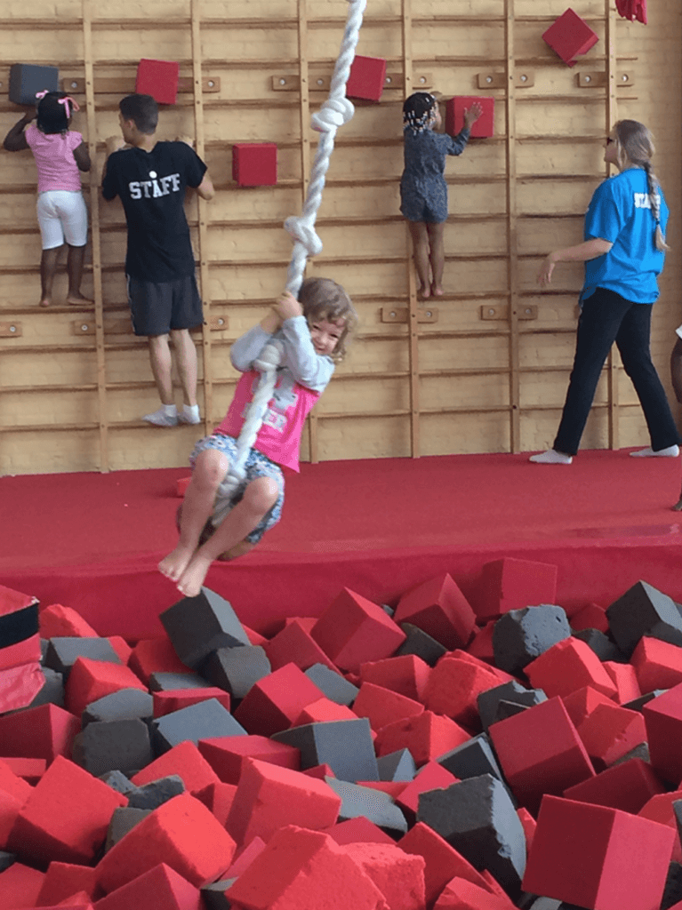 a girl swings from a rope over red and black foam blocks as kids balance on ledges in the wall behind her