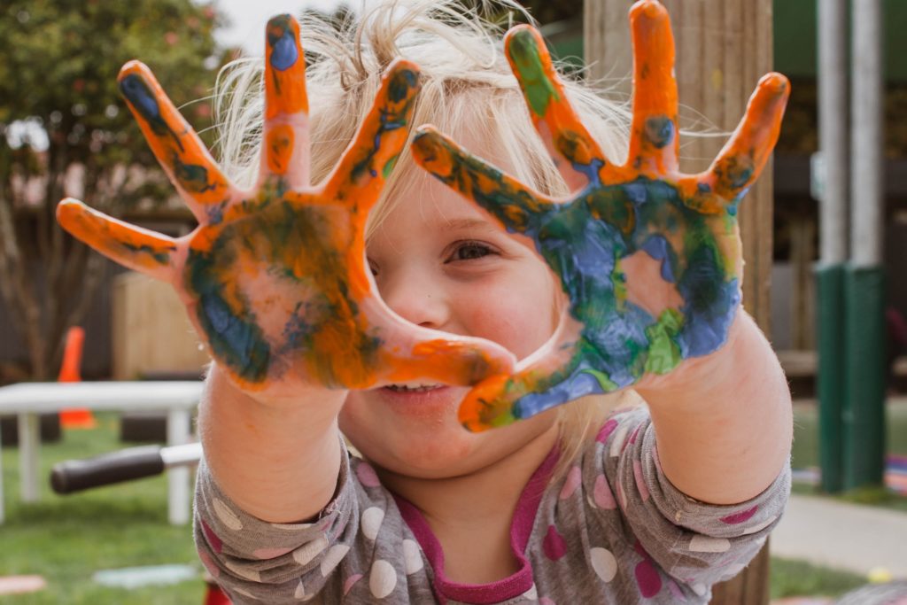 a girl smiles and puts out her hands, which are covered in orange, blue and green paint