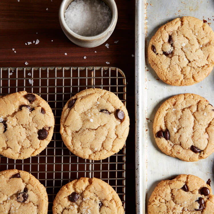 chocolate chip cookies sit on a cooling rack and tray with a bowl of salt nearby