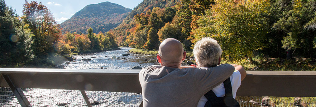 a man hugs a woman as they look out over the water surrounded by fall foliage and a mountain in the distance